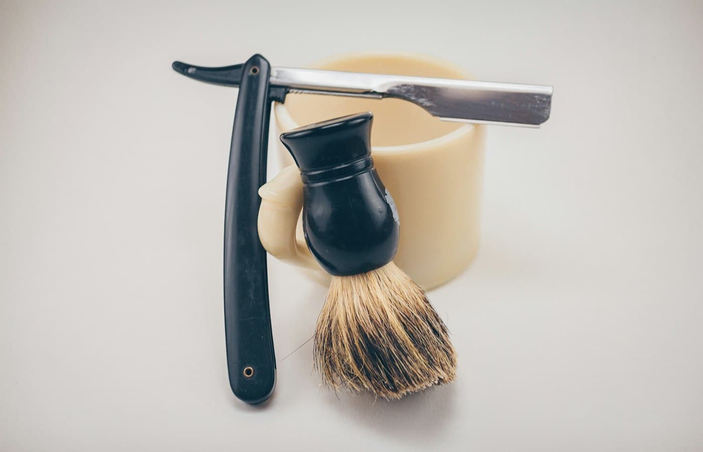 Black straight razor beside beige ceramic mug and shaving cream brush