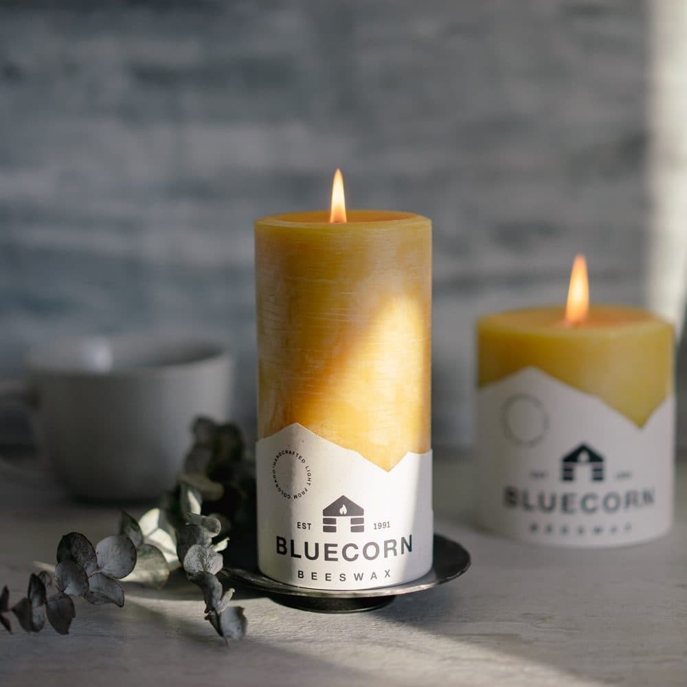 Two lit raw beeswax pillar candles with Bluecorn Beeswax labels on a dark tray beside eucalyptus