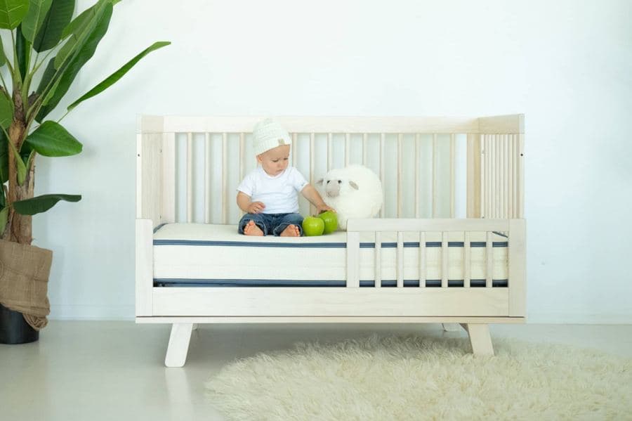 Baby sitting on a white crib mattress with blue piping inside a white wooden crib