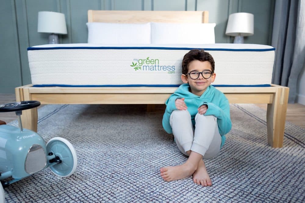 Boy sitting on the floor in front of a white My Green Mattress kids mattress on a wooden bed frame